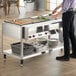 A stainless steel electric four pan hot food table with a lower shelf holding plates and bins, used for serving hot food buffet-style.