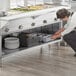 A woman in a white shirt and gray pants bending down to put food in a black pan on a steam table.