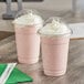 Two Choice clear plastic cups with pink liquid and white whipped cream with straws on a table.