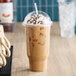 A large clear plastic cold cup with a dome lid, filled with iced coffee topped with whipped cream and chocolate syrup.