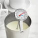 A stainless steel frothing thermometer clipped to a metal pitcher containing milk.