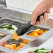 A hand using a Carlisle black plastic salad tong to pick up a piece of fruit from a food tray.