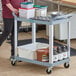 A woman pushing a gray Choice utility cart with two shelves holding food containers.