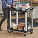 A woman pushing a black Choice utility cart with containers on it.