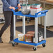 A woman pushing a blue Choice utility cart with containers on it.