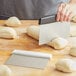 A person using a Choice stainless steel dough cutter with a black handle to cut dough on a cutting board.