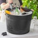 A woman pouring drinks into a Choice black beverage tub with wooden handles on a table outdoors.