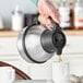 A person pouring coffee from a Bunn Zojirushi stainless steel coffee carafe into a white mug.