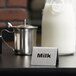 A Tablecraft stainless steel "Milk" tent sign in front of a metal pitcher of milk.