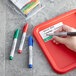 A hand holding a Dri Mark fine tip permanent marker and writing on a red tray.