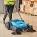 A woman using a blue and black Lavex outdoor sweeper in a grocery store aisle.