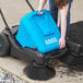 A woman using a Lavex blue manual sweeper to clean the ground.