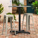 A Lancaster Table & Seating Excalibur outdoor table base with a plate and salt shaker on it and two white stools on a brick patio.