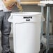 A man pouring food into a large white Rubbermaid ingredient storage bin.