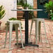 A black Lancaster Table & Seating Excalibur outdoor table base with white stools on a brick patio.