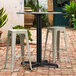 A Lancaster Table and Seating outdoor table base with a plate and salt and pepper shakers on it, with two white stools.