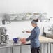 A woman in a blue uniform mixing ingredients in a large metal bowl on a white Baker's Lane shelf with clear plastic containers.