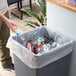 A man in green pants puts a plastic bottle in a Lavex trash bag in a trash can full of plastic bottles.