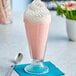 A glass of pink milkshake with whipped cream and a spoon on a white surface.