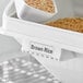 A white Baker's Mark ingredient bin with brown rice in it and a white spoon.