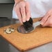 A hand using an Ateco chocolate shaver to peel a mushroom on a cutting board.