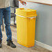 A man in blue jeans standing next to a yellow Lavex slim rectangular trash can with a plastic bag in it.