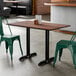 A rectangular standard height table with a textured walnut finish top and two black end base plates, shown with green metal chairs in a restaurant setting.