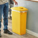 A man wearing jeans standing next to a yellow Lavex Slim Rectangular Recycle Bin with a bottle/can lid.