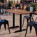 A rectangular standard height table with a textured Yukon oak finish and two black metal end base plates, set outdoors with black metal chairs.