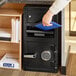 A person's hand holding a blue folder and opening a black steel depository safe.