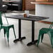 A rectangular standard height table with a smooth dark finish and two black end base plates, shown with green metal chairs in a restaurant setting.