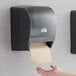 A hand reaching out to a Lavex Select black auto paper towel dispenser.