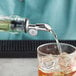 A person using a Tablecraft stainless steel liquor pourer to pour liquid into a glass on a counter.