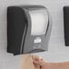 A hand putting a brown paper towel in a black Lavex Select paper towel dispenser on a wall.