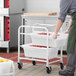 A man in a chef's uniform pushing a Regency mobile aluminum lug rack with white meat lugs full of food.