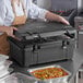 A person in a brown apron using a CaterGator black food pan carrier to hold a tray of food with rice and green onions.