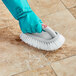 A gloved hand using a Quickie Contour All-Purpose Scrub Brush to clean a tile floor.