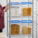 A woman in a red uniform standing next to a storage rack with Vigor clear polycarbonate food storage containers filled with pasta.
