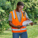 A man in a small/medium orange Ergodyne surveyor vest holding a clipboard and looking at a piece of paper.