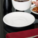 A white Tuxton china saucer holding a cup of coffee on a black table.
