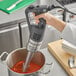 A hand using a Robot Coupe immersion blender to mix red sauce in a pot.