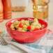 A close-up of an Acopa Capri Passion Fruit Red stoneware bowl filled with pasta, tomatoes, and olives on a cloth with a fork.