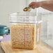A hand using a measuring spoon to pour pasta into a large clear plastic container.