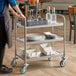 A three-shelf stainless steel utility cart holding glassware, bottles, plates, and trays in a restaurant setting.