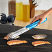 A hand holding a Choice blue stainless steel scalloped tong over a grill with food on it.