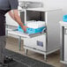 A man putting a blue tray of dishes into a Noble Warewashing undercounter dishwasher on a stand.