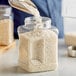 A person pouring white rice into a 48 oz. square plastic jar.