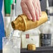 A hand using a Franmara gold-plated cocktail shaker to pour a drink into a glass.