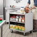 A man using a Regency stainless steel utility cart to transport food in a white container with a blue lid.