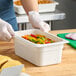 A person in gloves holding a white Cambro food container filled with colorful peppers.